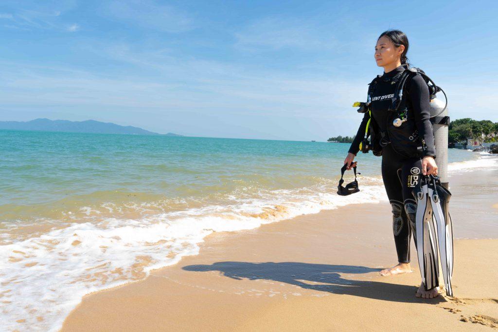 A woman in a wetsuit stands on a sandy beach, holding diving fins and a mask, with the ocean and blue sky in the background. She appears ready for scuba diving.